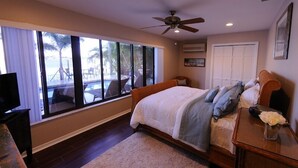 A bedroom with a large bed, a ceiling fan, a TV, and a view of a pool and palm trees. - Pasa Tiempo Private Waterfront Resort - Adults Only (St. Pete Beach)