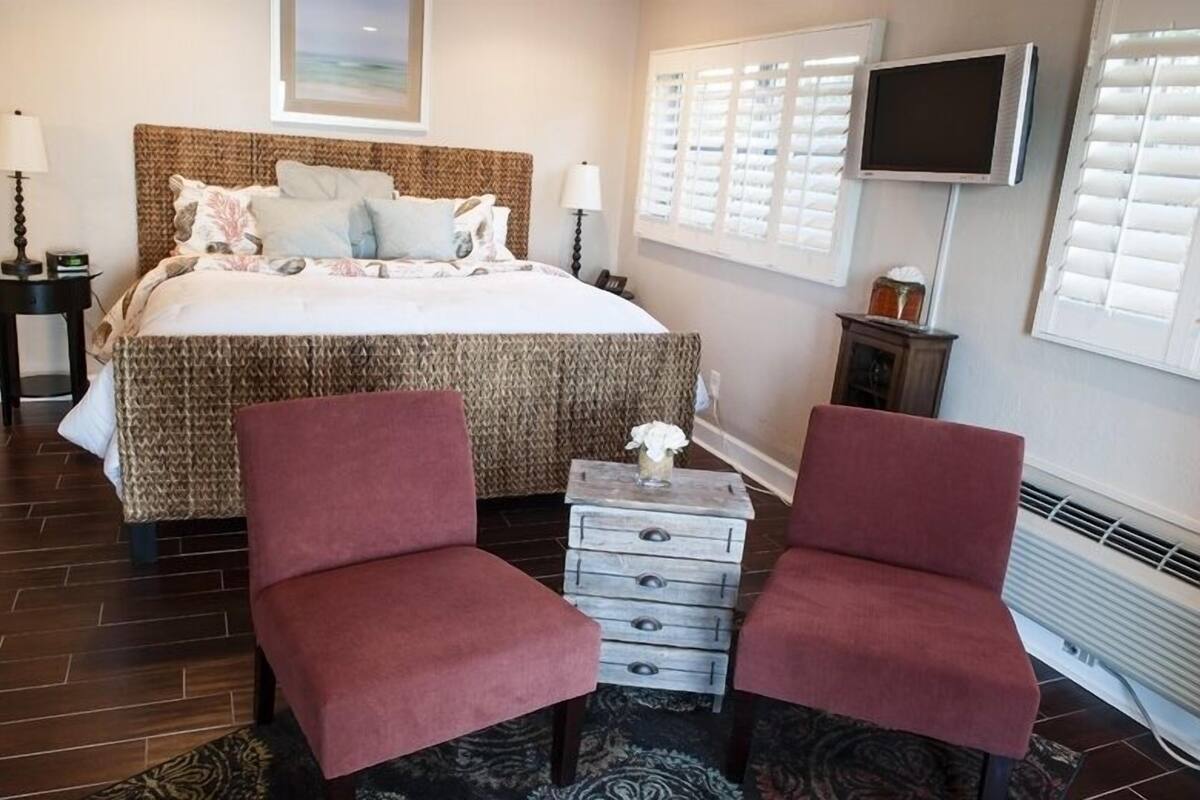 A bedroom with a large bed, two red chairs, a small side table, a TV, and a window with blinds.
