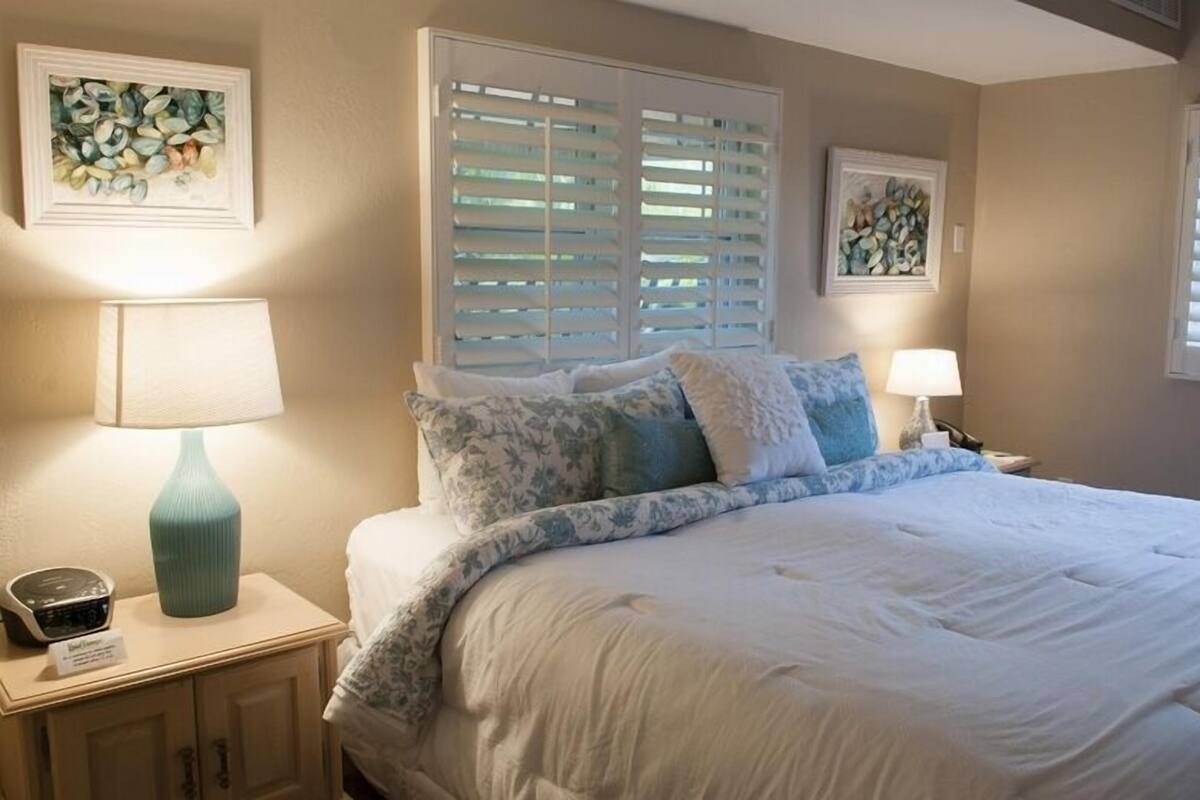 A bedroom with a bed, two bedside tables, a lamp, a radio, and artwork on the walls.