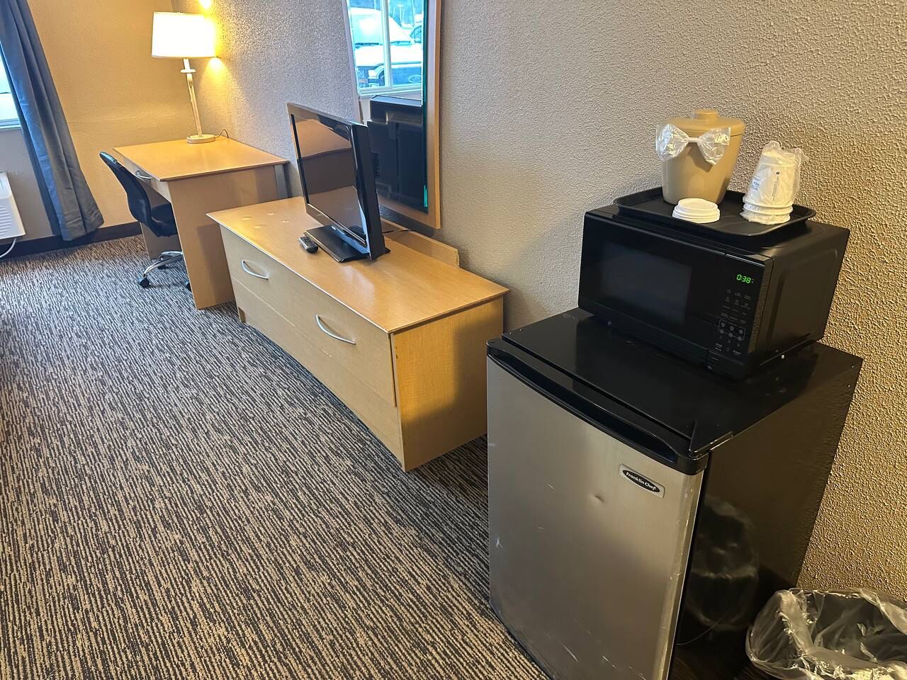 Habitación de hotel compacta con escritorio, refrigerador pequeño, microondas y un cesto de basura.