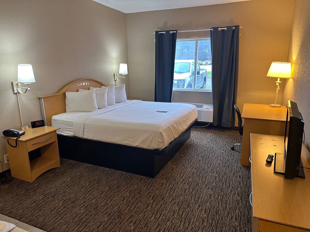 A hotel room with a single bed, bedside tables, a desk with a computer, and a window with curtains.