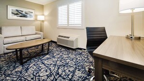 A hotel room with a sofa, a small table, a lamp, a chair, and a window with blinds. - Quality Suites Houston Hobby Airport (Houston)