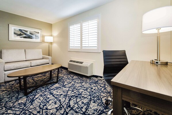 A hotel room with a sofa, a small table, a lamp, a chair, and a window with blinds.
