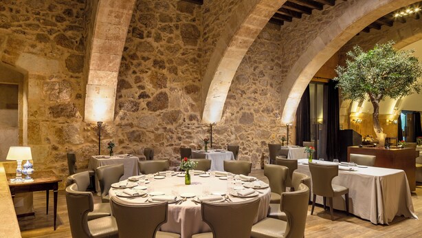 A stone-walled dining area with round tables and chairs, wooden beams on the ceiling, and a large tree in the background.