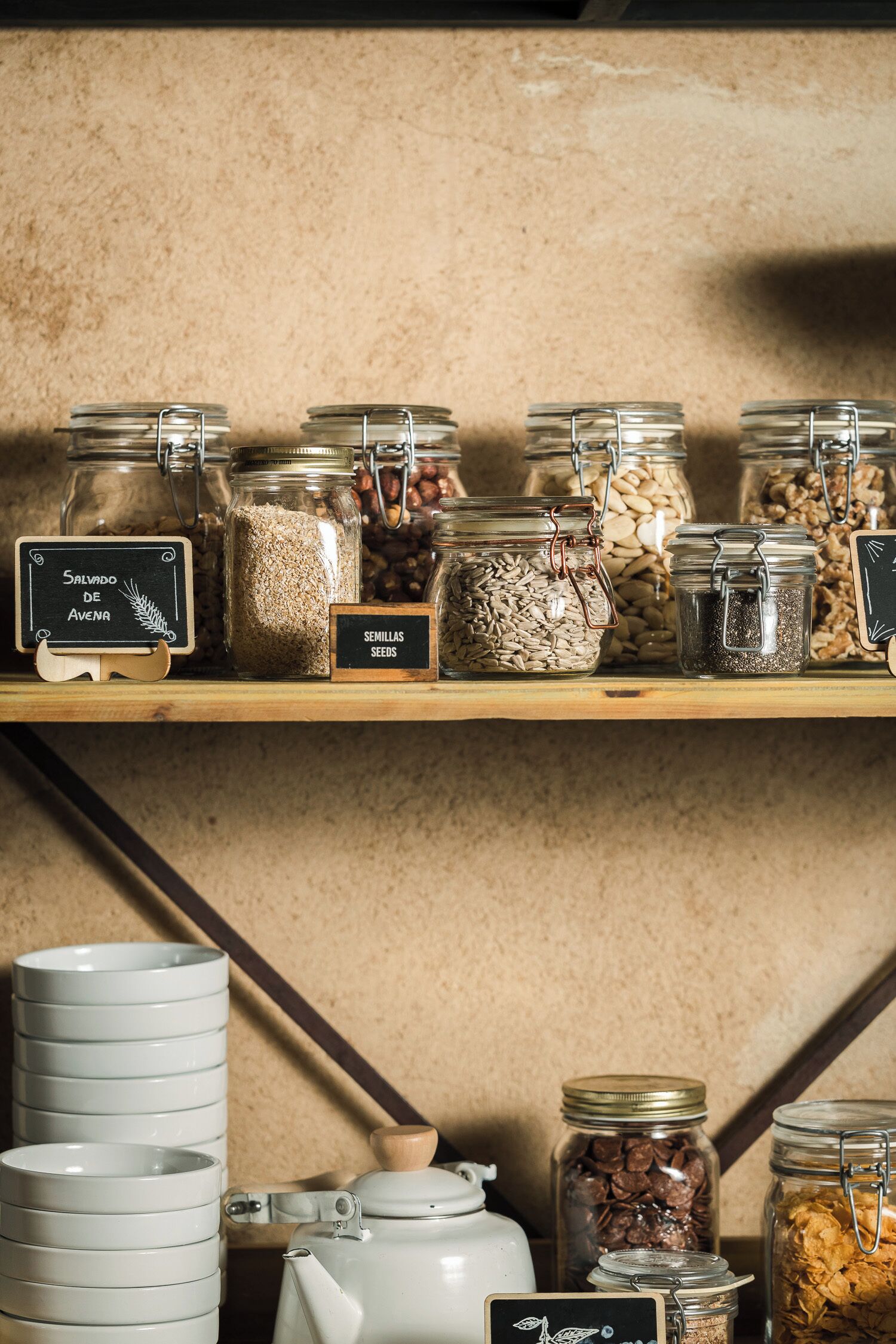 Una estantería con frascos de cereales y semillas, un cartel de pizarra y platos blancos apilados.