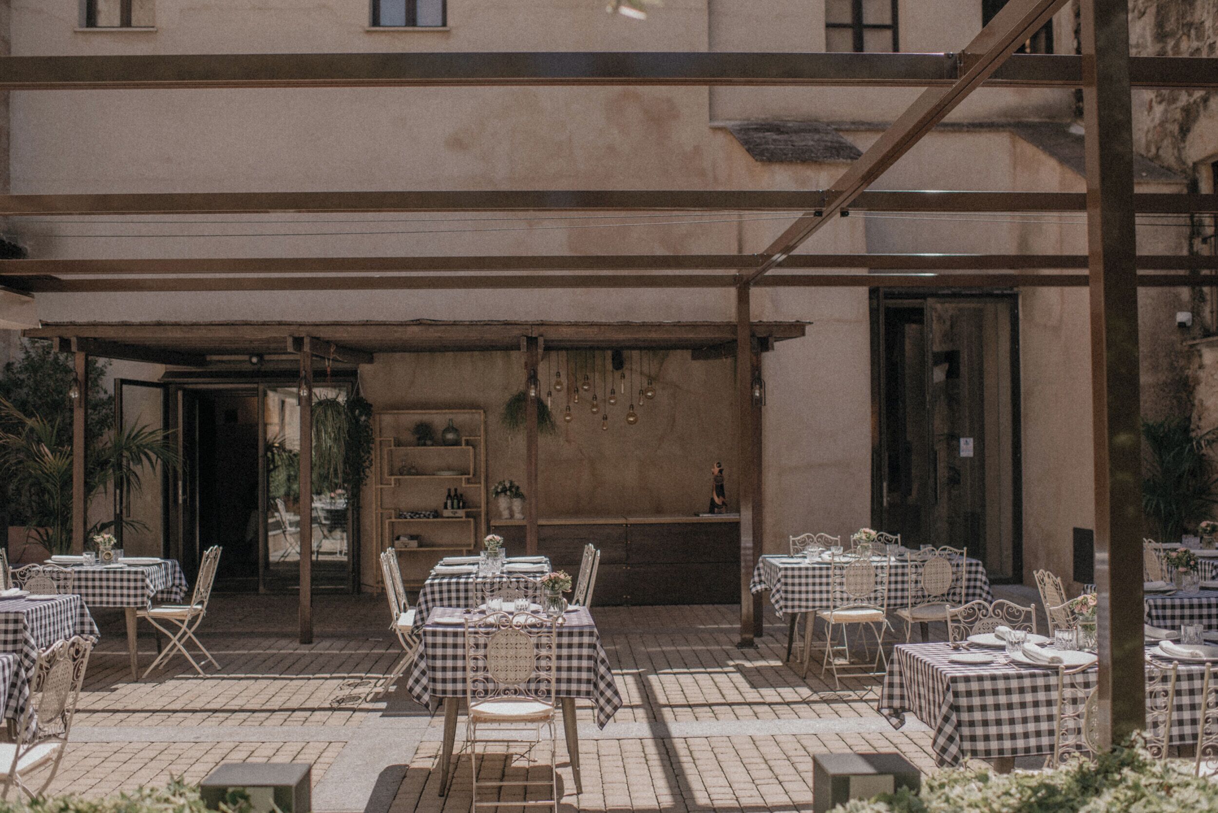 An outdoor patio with checkered tablecloths and metal chairs, surrounded by a building with a rustic appearance.