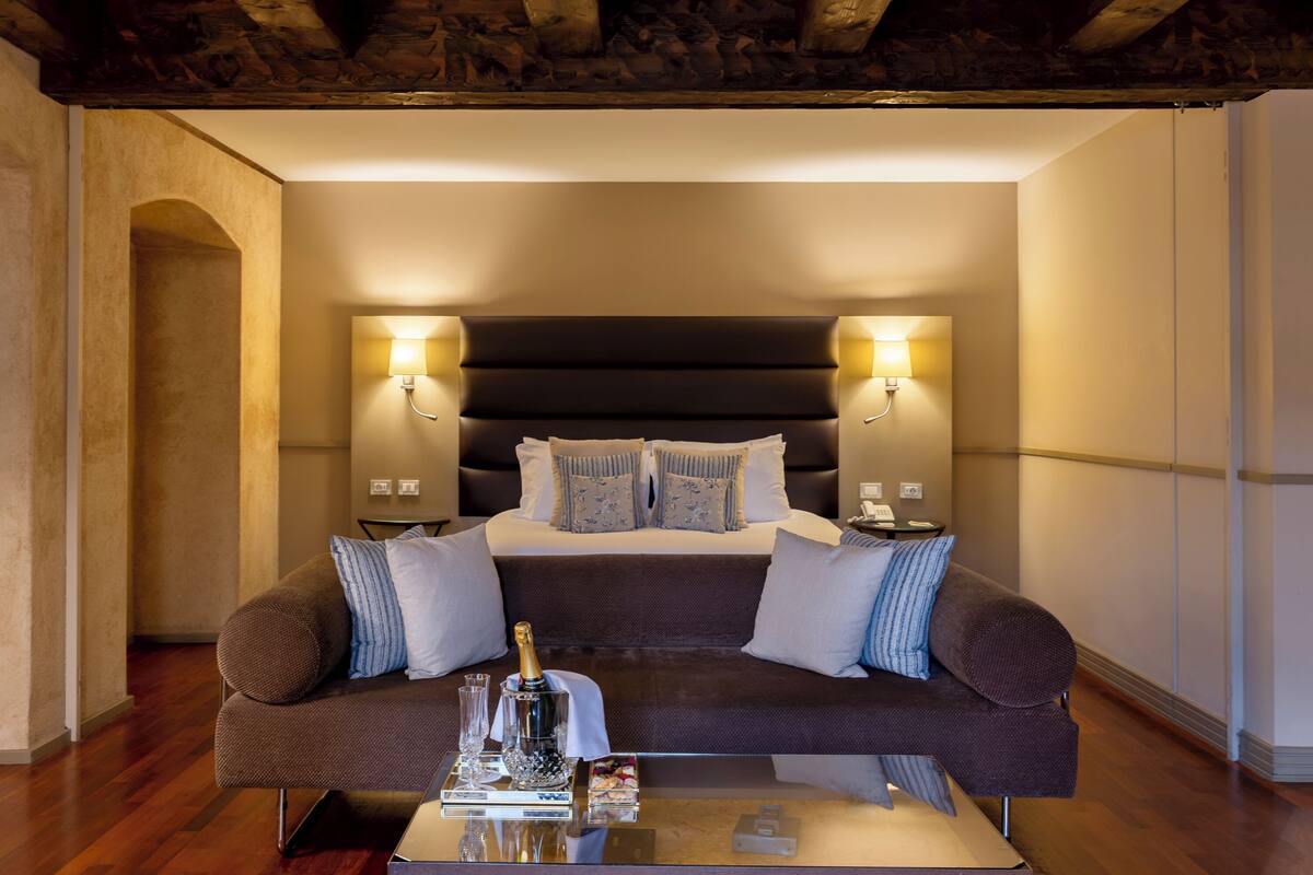 Een moderne hotelkamer met een groot bed, twee bedlampen, een bruine bank en een glazen salontafel met een fles en glazen.