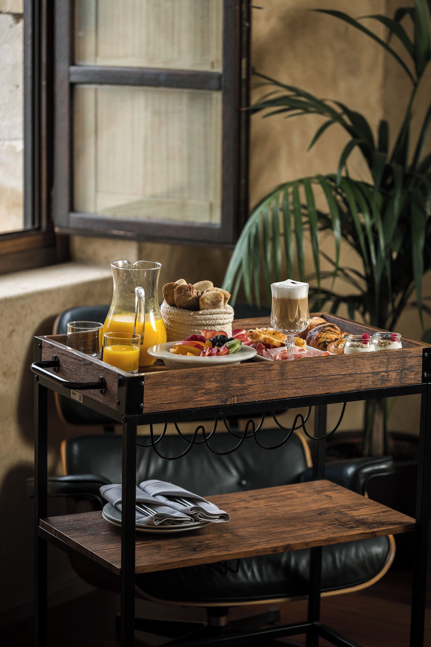 A breakfast tray with orange juice, coffee, pastries, and a croissant on a wooden table in a room with a window and a plant.
