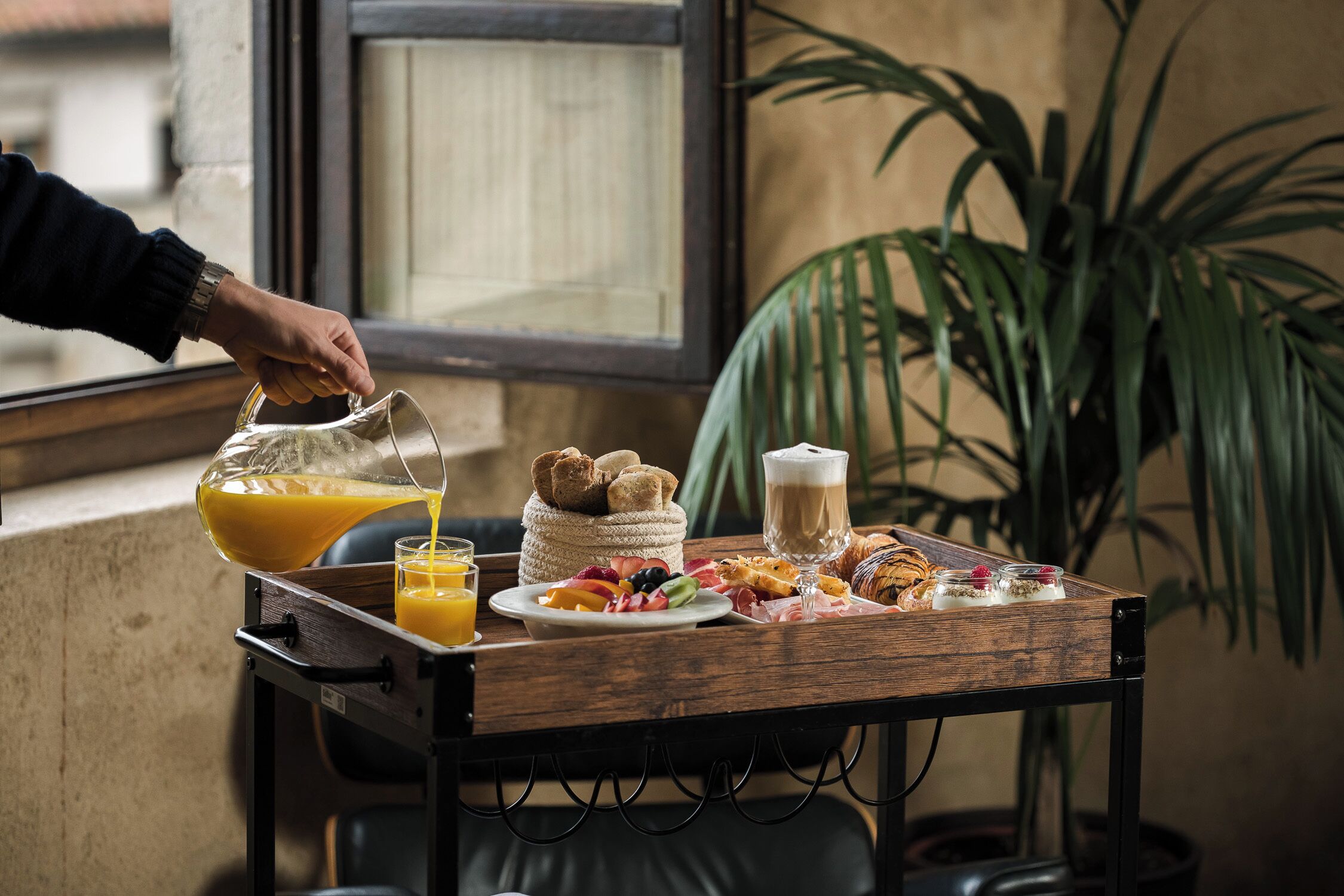 Una bandeja de desayuno con jugo de naranja, croissants y una taza de café sobre una mesa de madera cerca de una ventana.