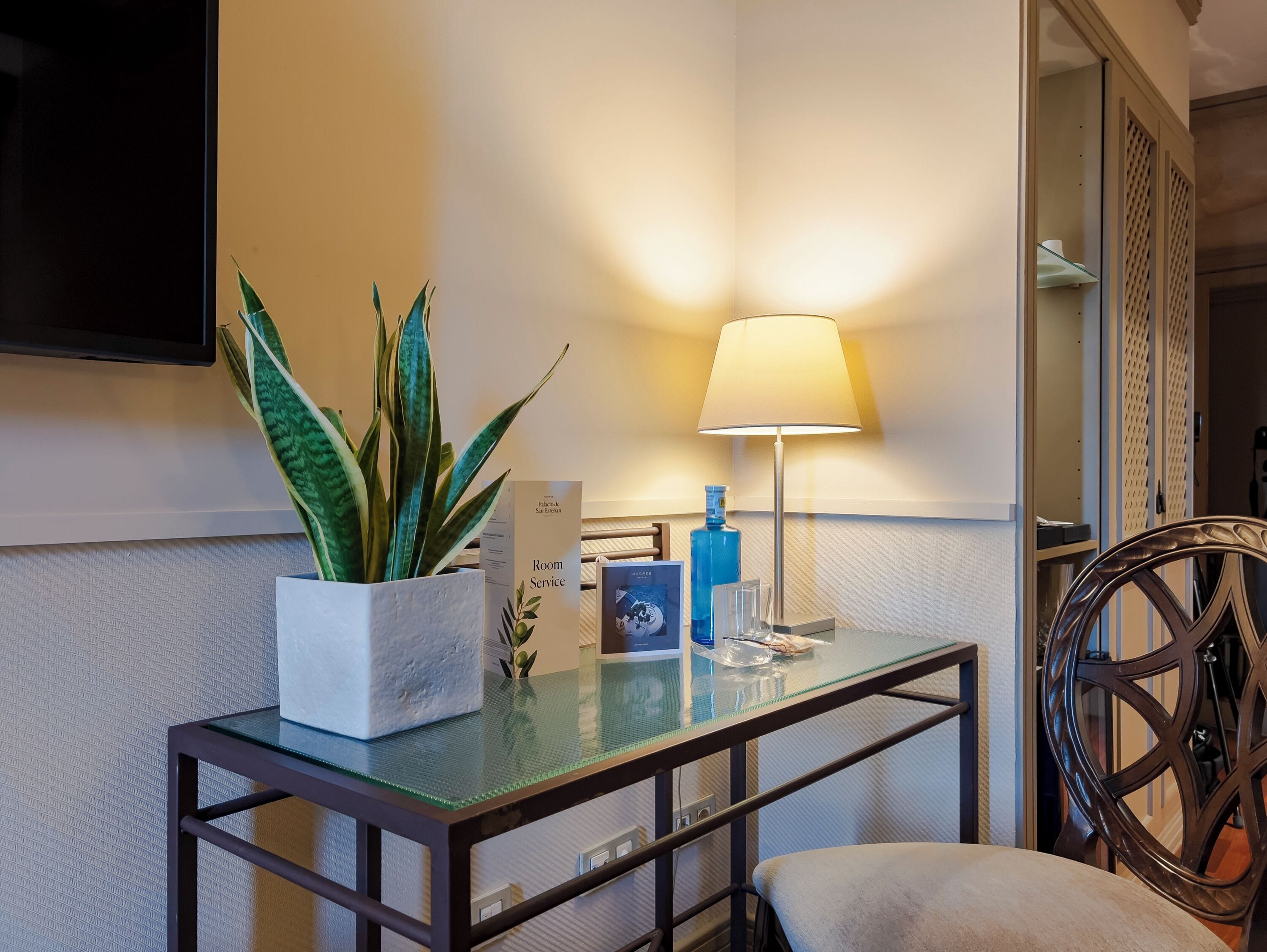 Una habitación de hotel con una mesa de vidrio, una planta en maceta, una lámpara y una silla.