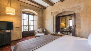 A bedroom with a large bed, a television, a chair, a table with a tray of items, and a view through an arched doorway.
