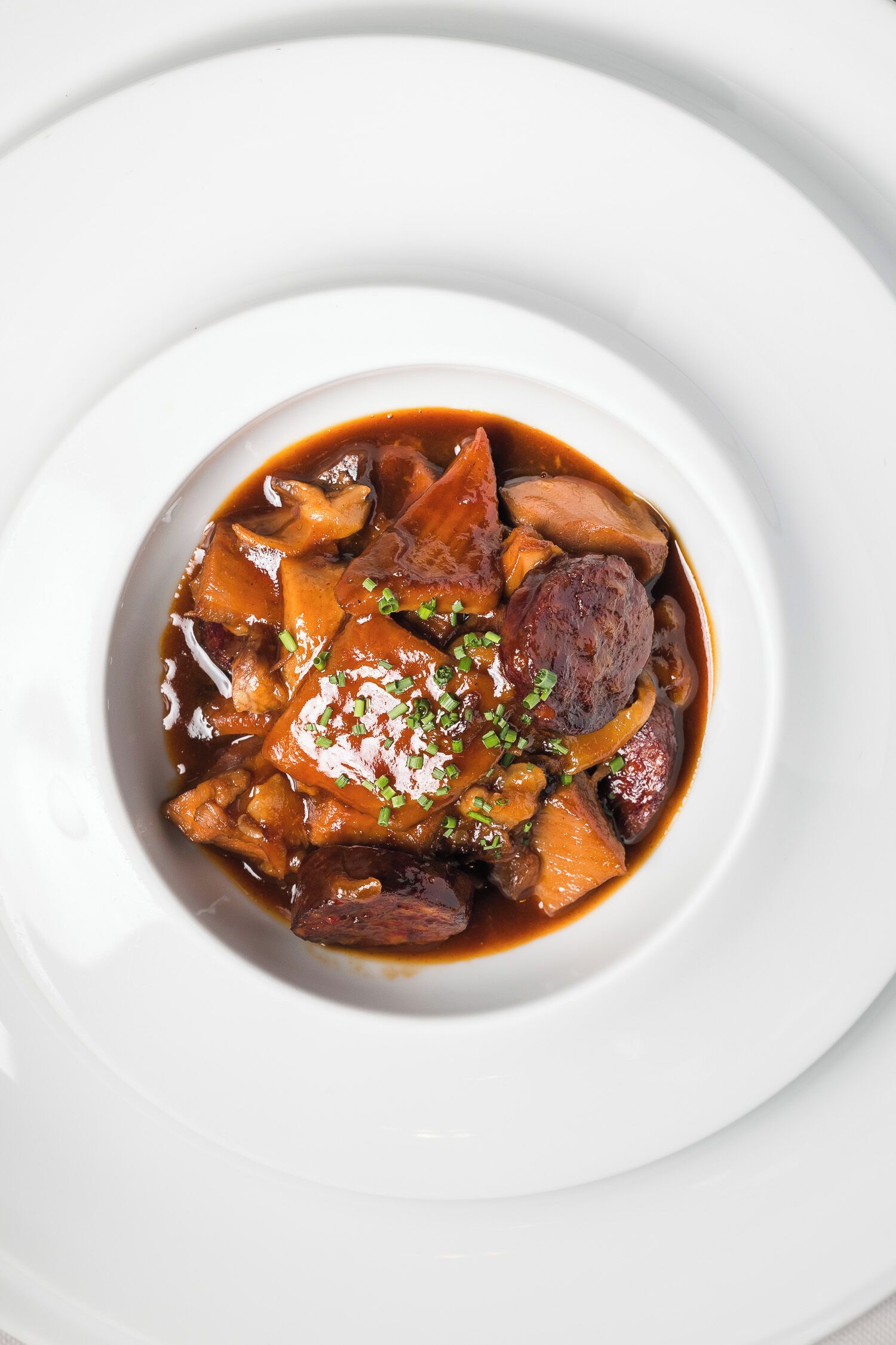 Un plato blanco con guiso de carne y verduras en salsa marrón, adornado con hierbas verdes picadas.