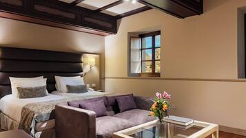 A hotel room with a large bed, a sofa, a glass coffee table, and a window with a view of buildings.