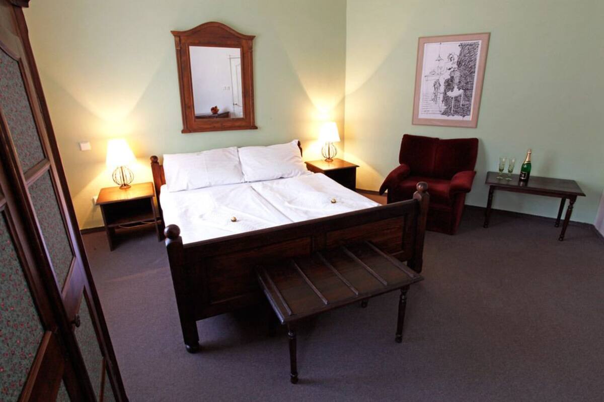 A bedroom with a wooden bed, two bedside tables with lamps, a red armchair, and a small table with glasses and a bottle.