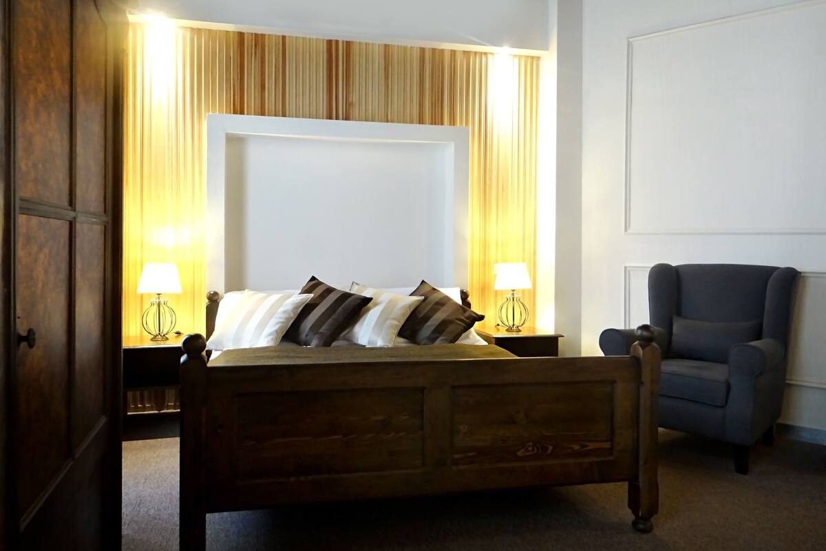 A bedroom with a wooden bed, two bedside lamps, a chair, and a large window with curtains.