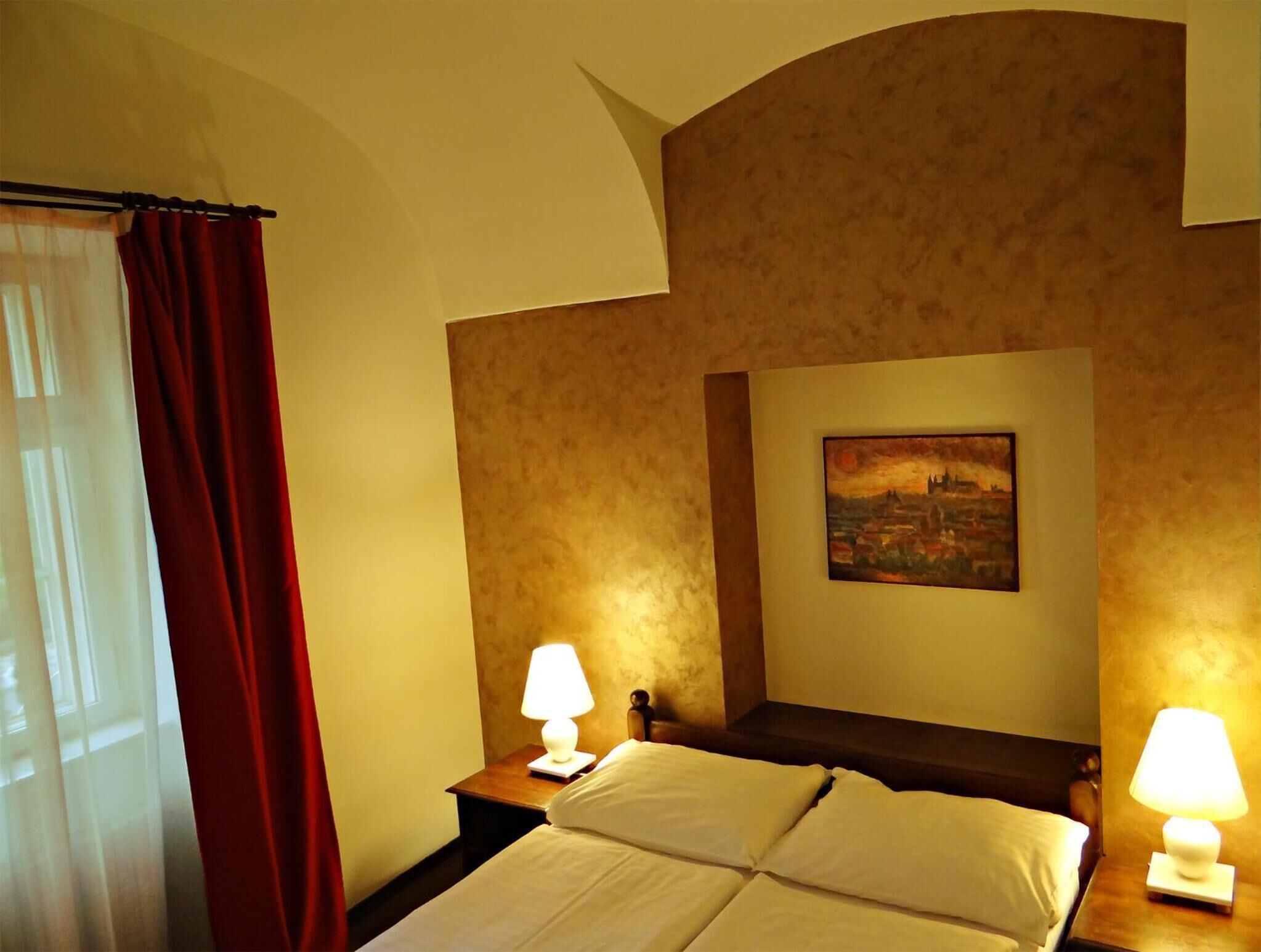 A hotel room with a bed, two bedside lamps, a painting on the wall, and a red curtain.