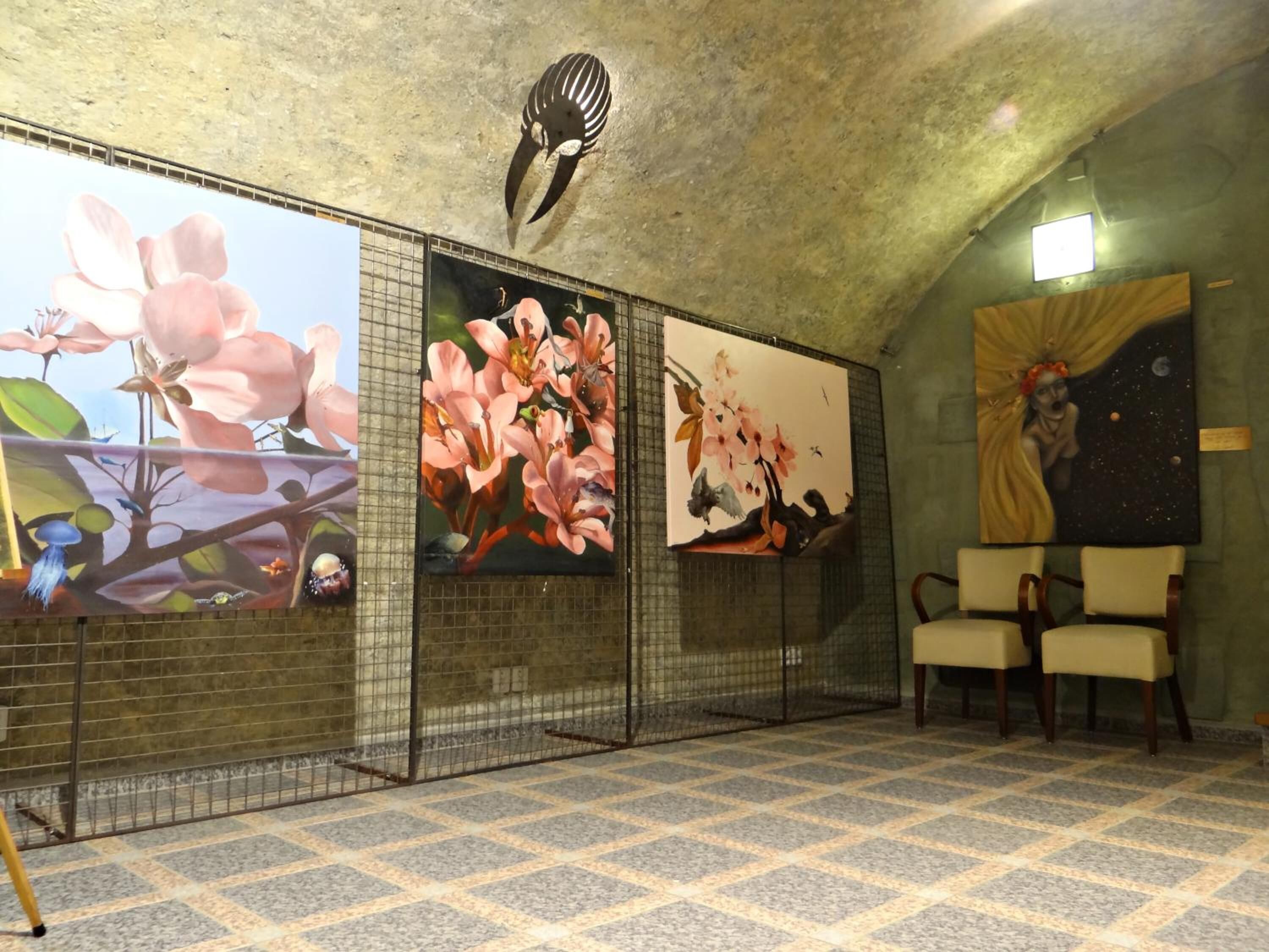 Um quarto com soalho ladrilhado, parede com três grandes pinturas florais e teto com um mural de aves, e duas cadeiras.