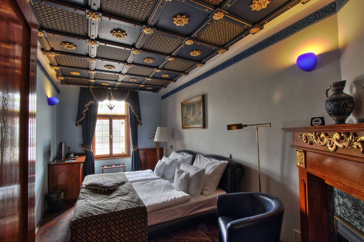 A room with a bed, a chair, a desk, a lamp, a fireplace, and a window.
