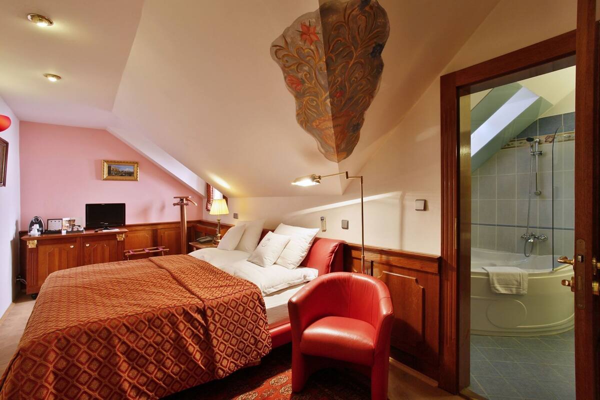 A hotel room with a bed, a red chair, a desk with a computer, a TV, and a bathroom with a bathtub.