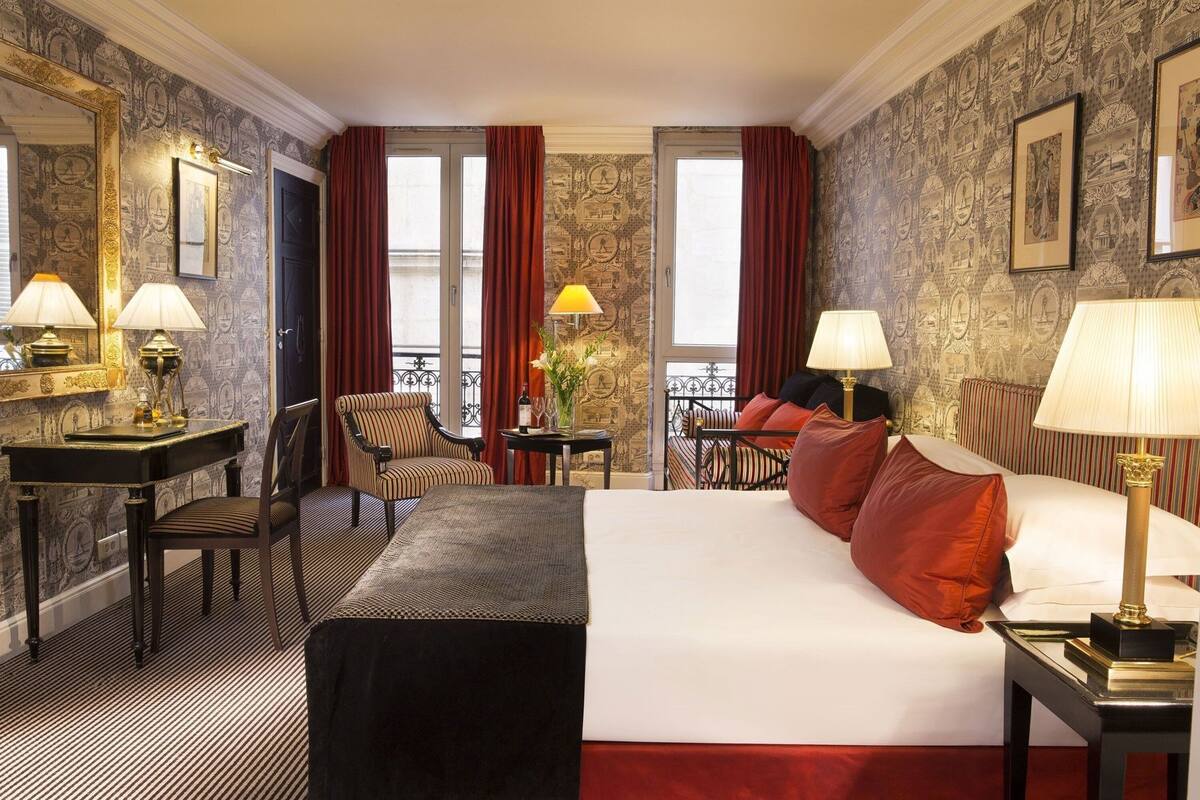 Hotelkamer met een groot bed, twee fauteuils, een kleine tafel, een bureau en een balkon met gordijnen.