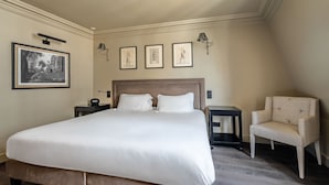 Un lit bien fait, avec une tête de lit, deux tables de chevet, une chaise et des œuvres d’art encadrées accrochées au mur.