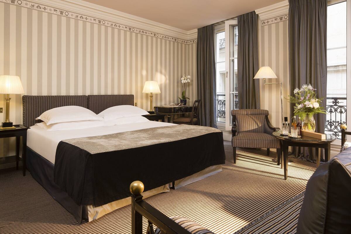 Hotelkamer met een groot bed, twee nachtkastjes met lampen, een stoel, een klein tafeltje met een vaas, en uitzicht op de straat door een raam met gordijnen.