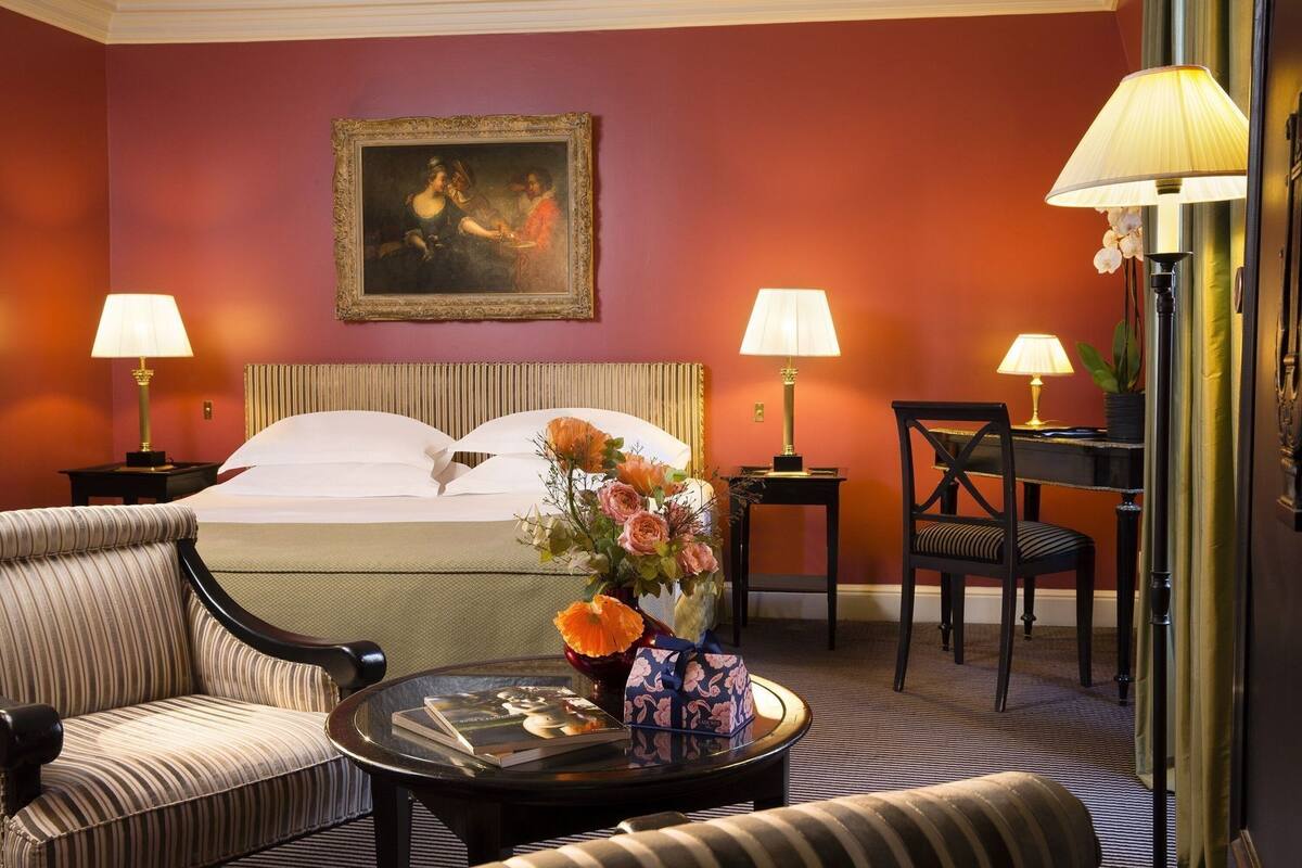 Een hotelkamer met een bed, twee stoelen, een bureau, een lamp, een vaas met bloemen en een schilderij aan de muur.
