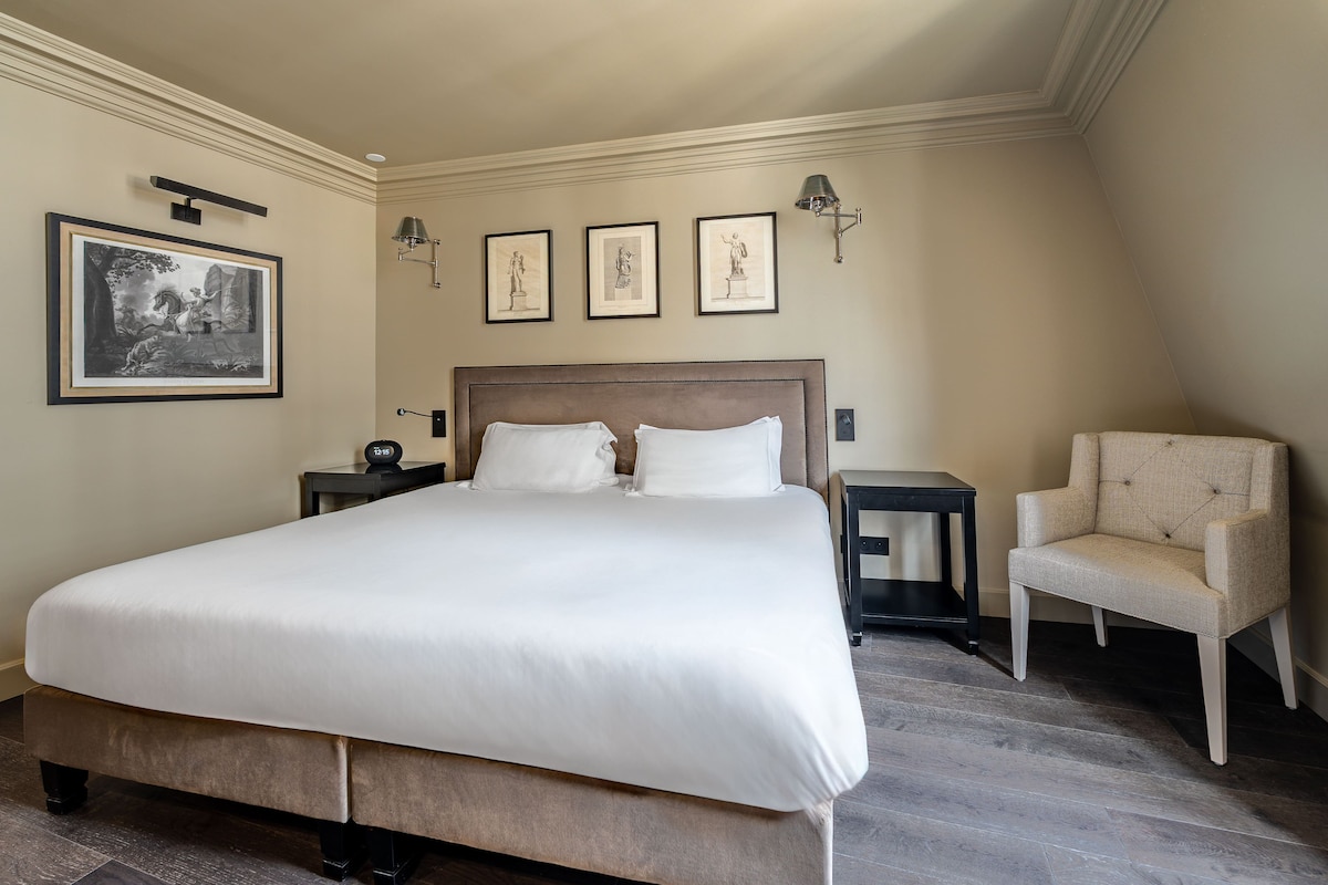 A neatly made bed with a headboard, two bedside tables, a chair, and framed artwork on the wall.