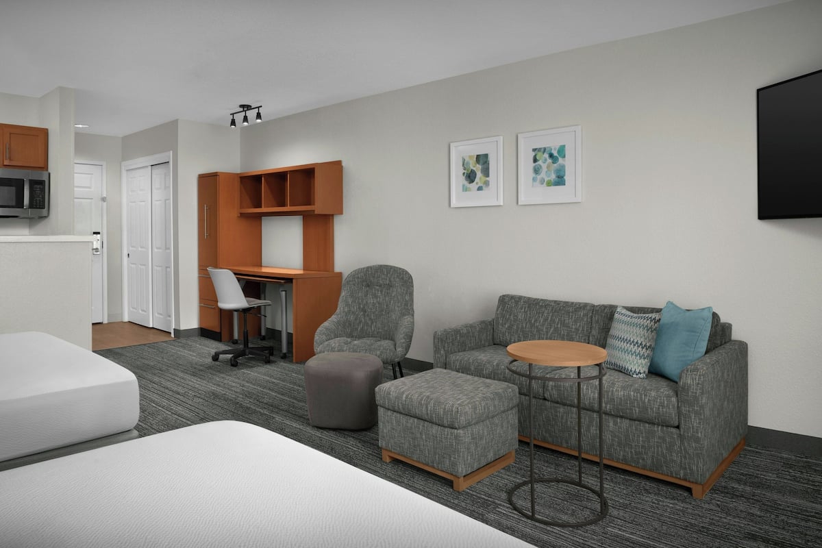 Ein modernes Hotelzimmer mit grauem Sofa, einem kleinen runden Tisch, einem grauen Sessel, einem weißen Bett, einem Schreibtisch mit Stuhl und einem an der Wand befestigten Fernseher.