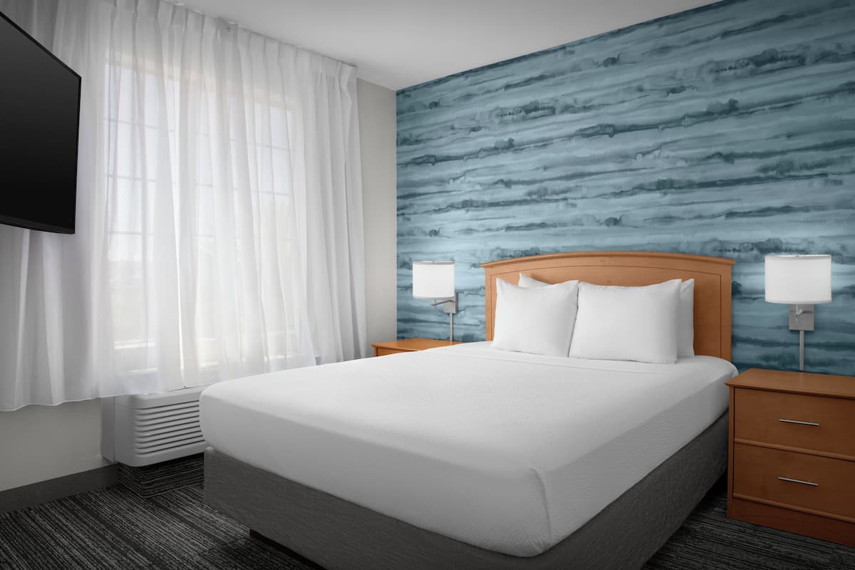 Ein Hotelzimmer mit einem großen Bett, zwei Nachttischen mit Lampen, einem an der Wand montierten Fernseher und einem Fenster mit durchsichtigen Vorhängen.