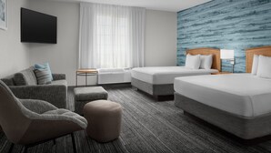 A hotel room with a flat-screen TV, two beds, a sofa, an ottoman, a window with sheer curtains, and a blue textured wall. - Towneplace Suites Abq Airport (Albuquerque)