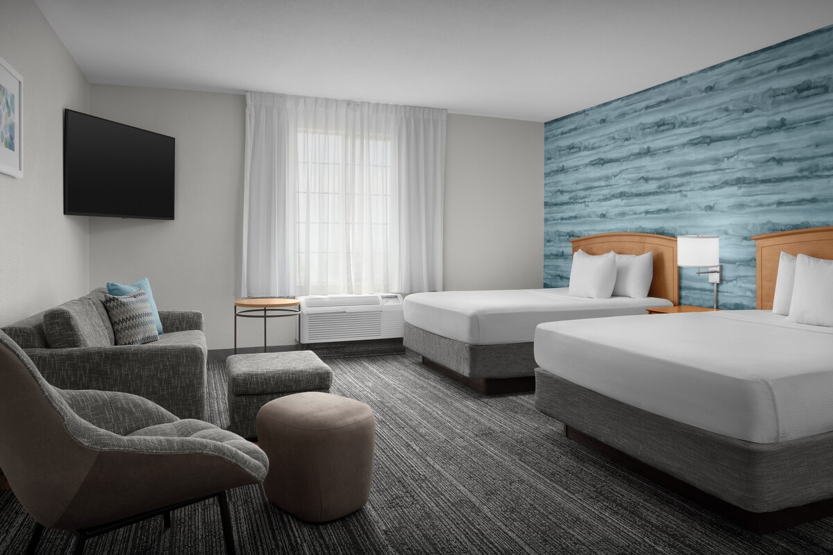 Habitación de hotel con televisión de pantalla plana, dos camas, un sofá, un reposapiés, una ventana con cortinas traslúcidas y una pared azul con textura.