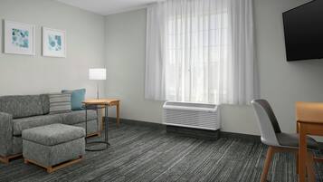 Una habitación de hotel moderna con un sofá gris, una mesita redonda pequeña, una silla, un televisor de pantalla plana y un ventanal con cortinas translúcidas.