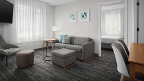 A modern hotel room with a grey sofa, ottoman, and armchair, a flat-screen TV, and two abstract art pieces on the wall. - Towneplace Suites Abq Airport (Albuquerque)