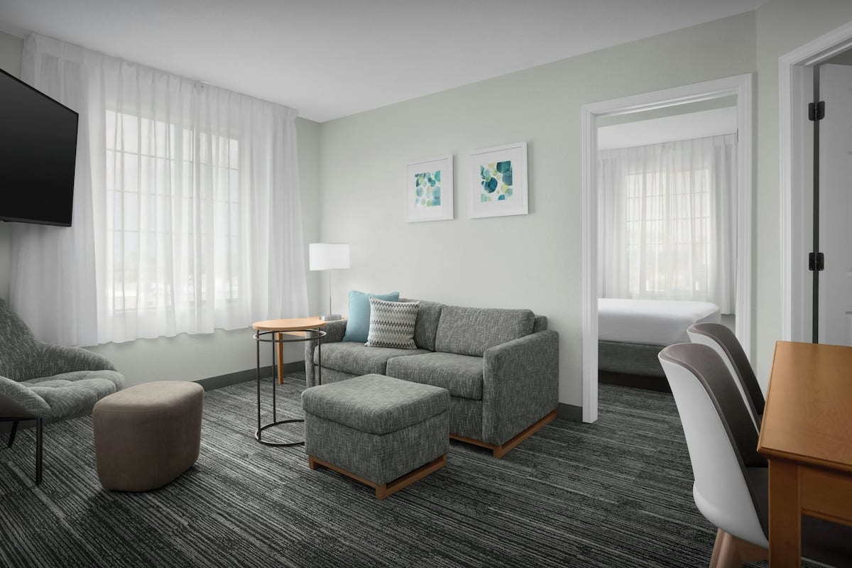 Ein modernes Hotelzimmer mit grauem Sofa, Ottoman und einem kleinen Tisch mit Lampe. An der Wand befinden sich zwei gerahmte Bilder und ein montierter Fernseher.