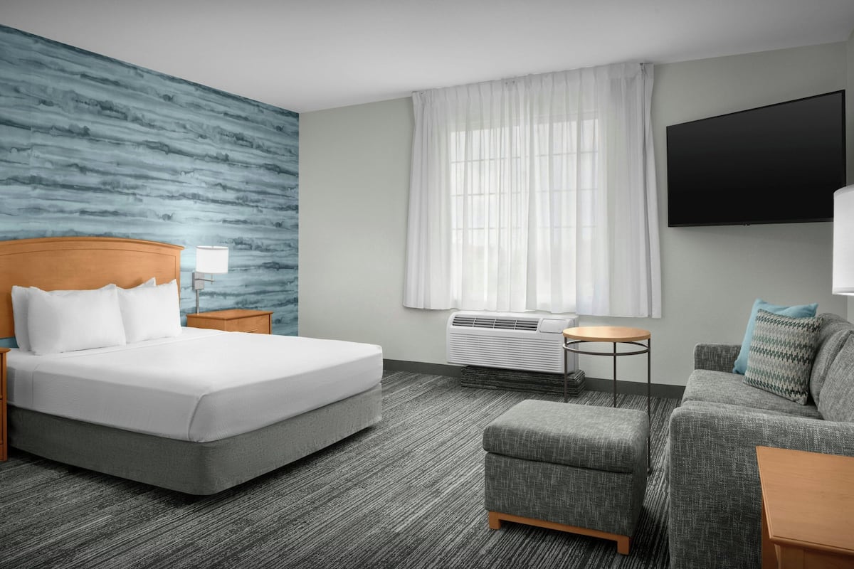 Ein Hotelzimmer mit einem großen Bett, einem grauen Polsofa, einem kleinen Hocker, einem Flachbildfernseher, einem Fenster mit weißen Vorhängen und einer Klimaanlage.