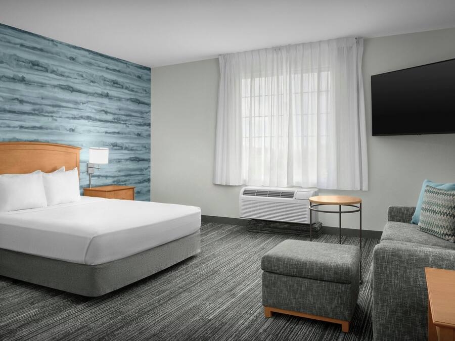 A hotel room with a large bed, a grey upholstered sofa, a small ottoman, a flat-screen TV, a window with white curtains, and an air conditioning unit.