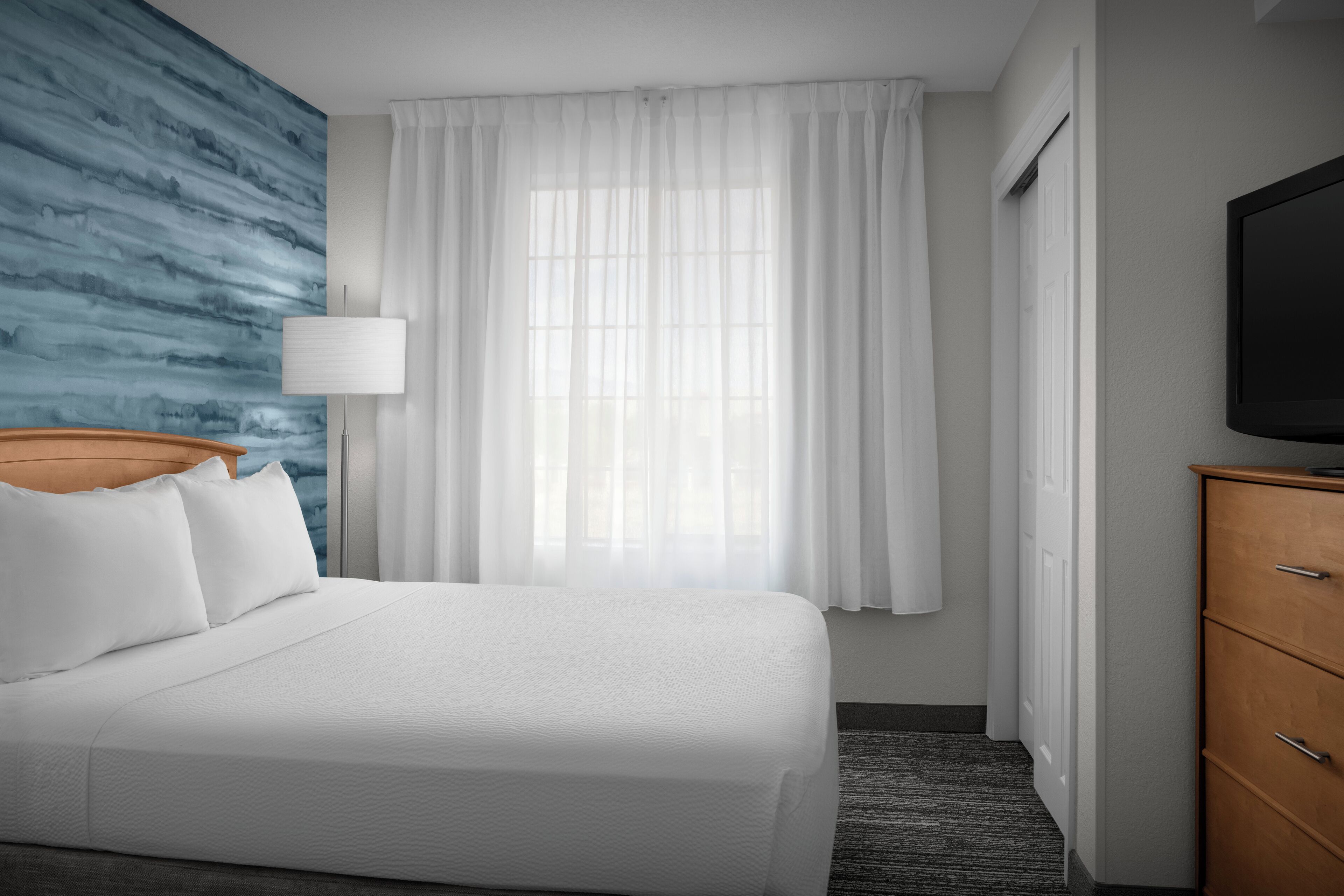 Quarto de hotel com cama, televisão, luminária e janela com cortinas.