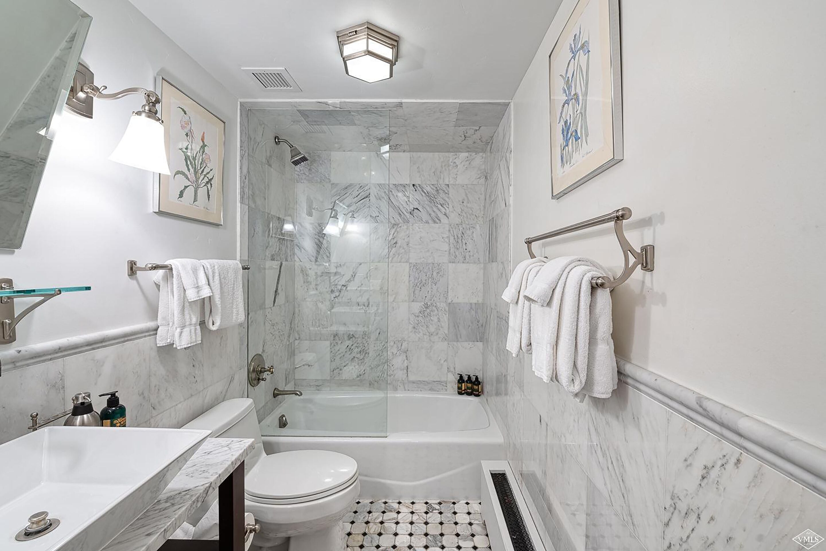 Une salle de bain moderne avec un lavabo, des toilettes et une baignoire en marbre. Les murs sont ornés de tableaux encadrés et il y a une douche avec une porte en verre.