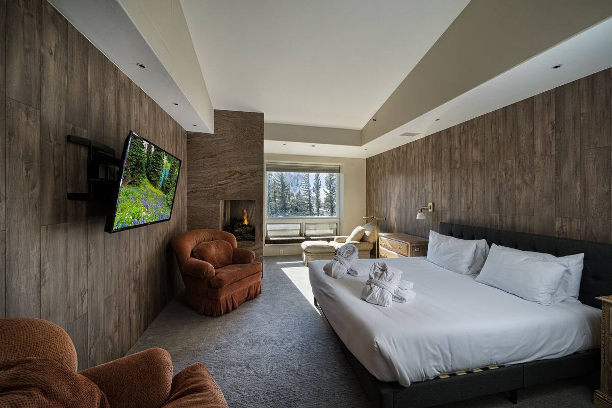 A modern hotel room with a large bed, a flat-screen TV mounted on the wall, a fireplace, and a view of trees through the window.