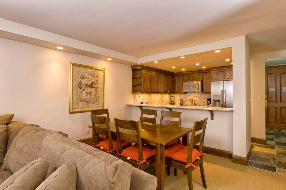 A dining area with a wooden table and chairs, a kitchen with wooden cabinets and stainless steel appliances, and a framed picture on the wall.