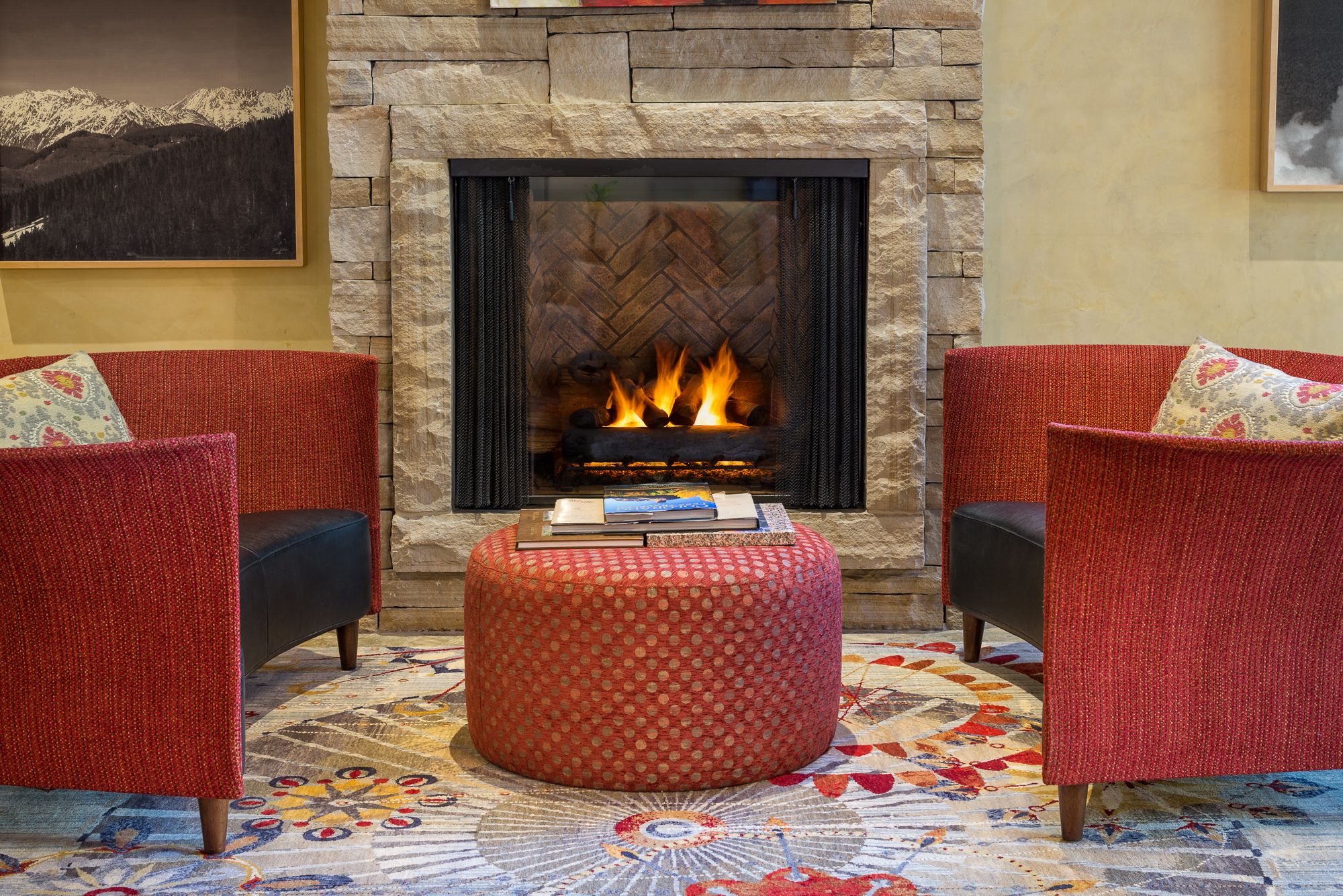 Une pièce chaleureuse avec une cheminée en pierre, deux fauteuils rouges à motifs, un pouf à motifs et un tapis coloré.