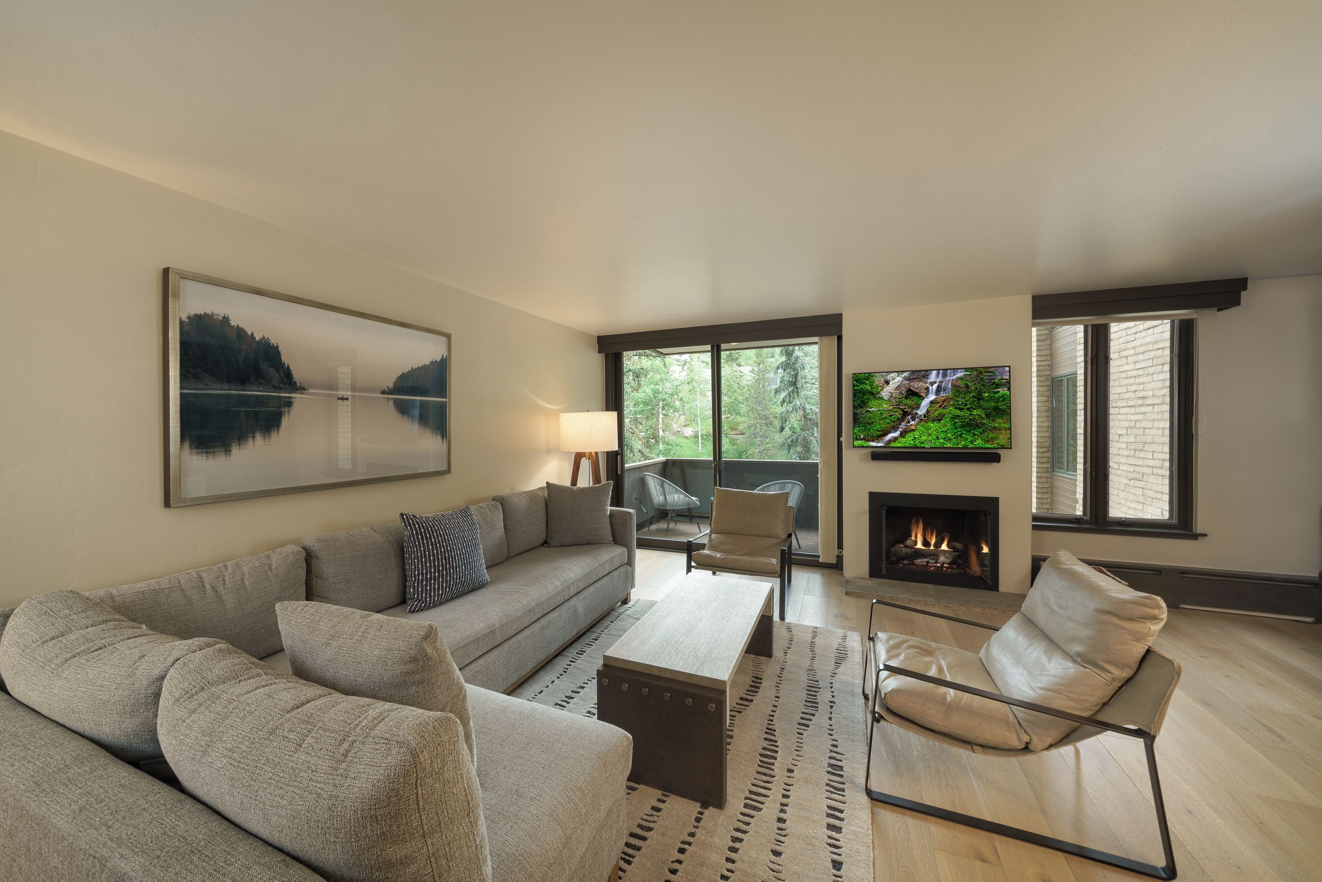 A modern living room with a sofa, coffee table, and a fireplace. There is a television mounted on the wall and a window with a view of greenery.
