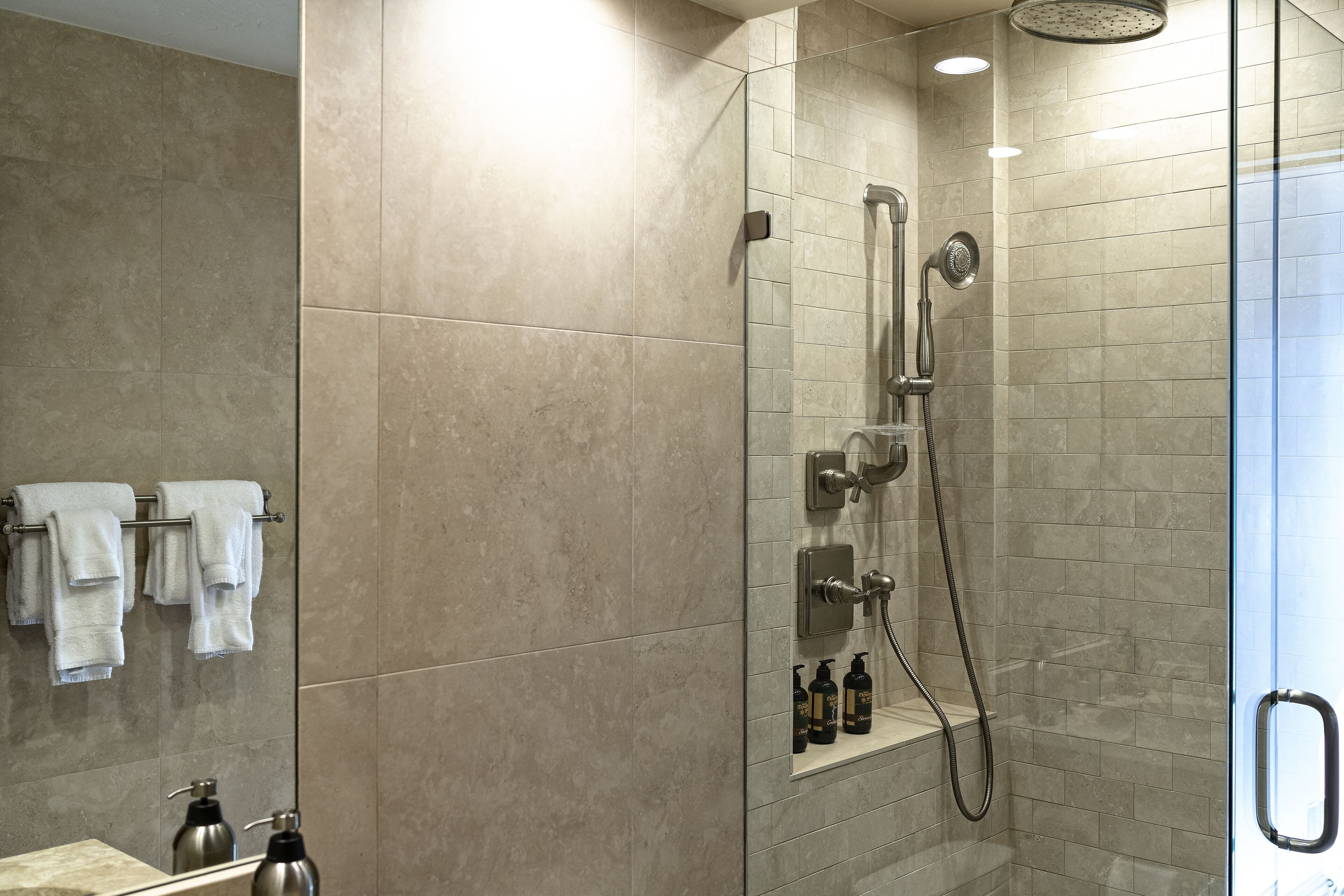 A modern bathroom with a glass-enclosed shower, beige tiles, and a shelf with toiletries.