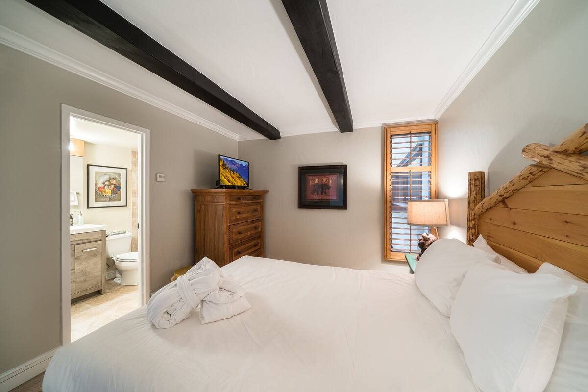 A bedroom with a large bed, wooden headboard, a dresser, a television, and a window with blinds.