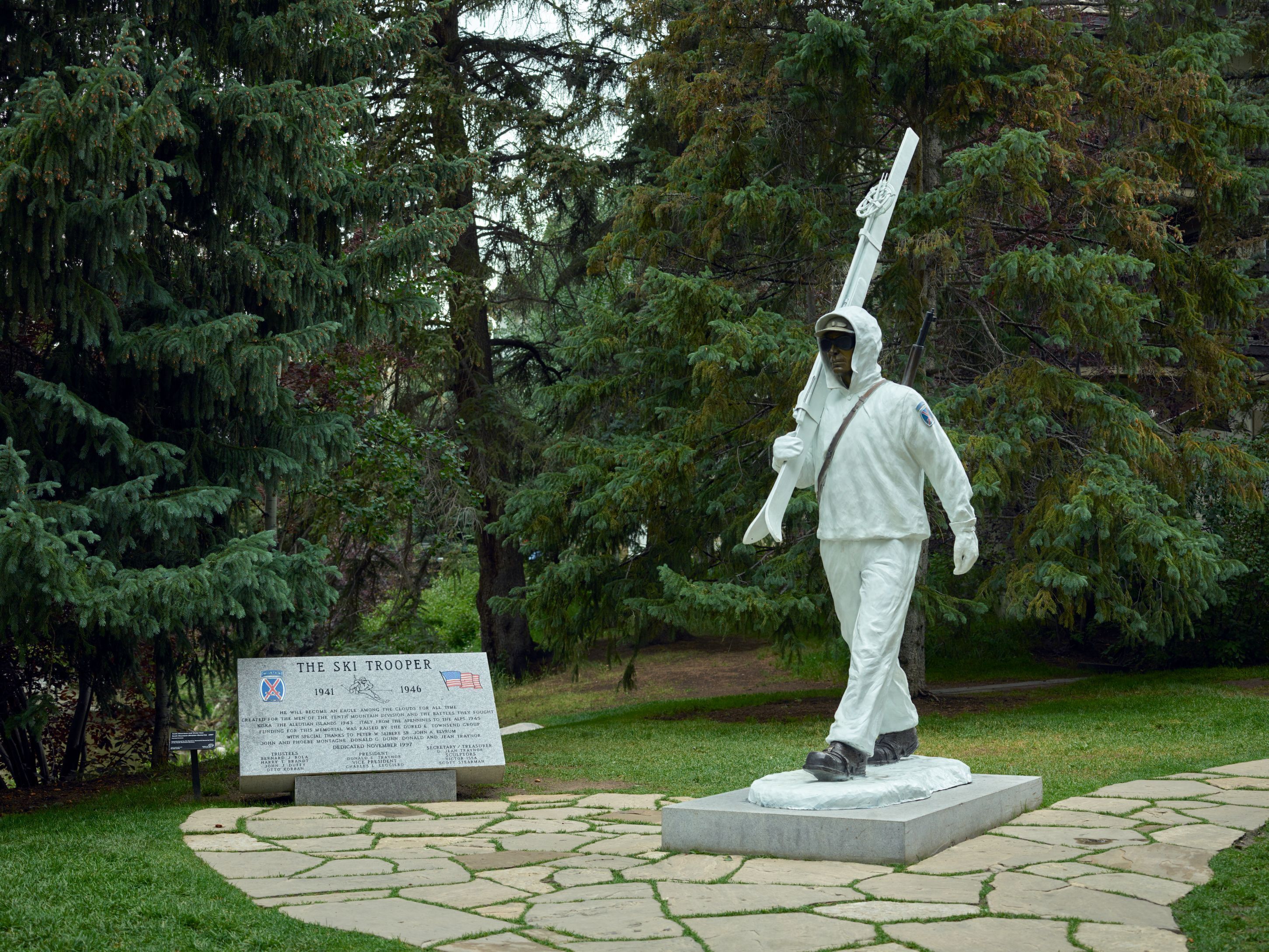 Eine Statue eines Ski-Truppiers in einem Park mit einer Gedenktafel, umgeben von Bäumen.