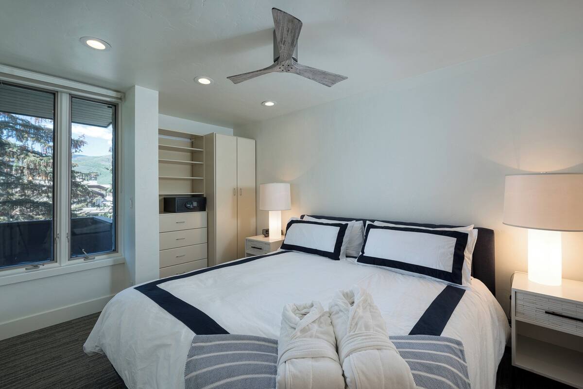 A hotel room with a large bed, a ceiling fan, a window with a view, a bedside table with a lamp, and a wardrobe.