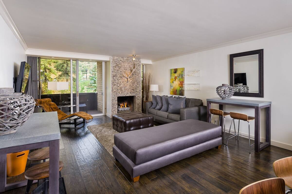 A modern living room with a fireplace, a sofa, a dining area, and a view of the outdoors.