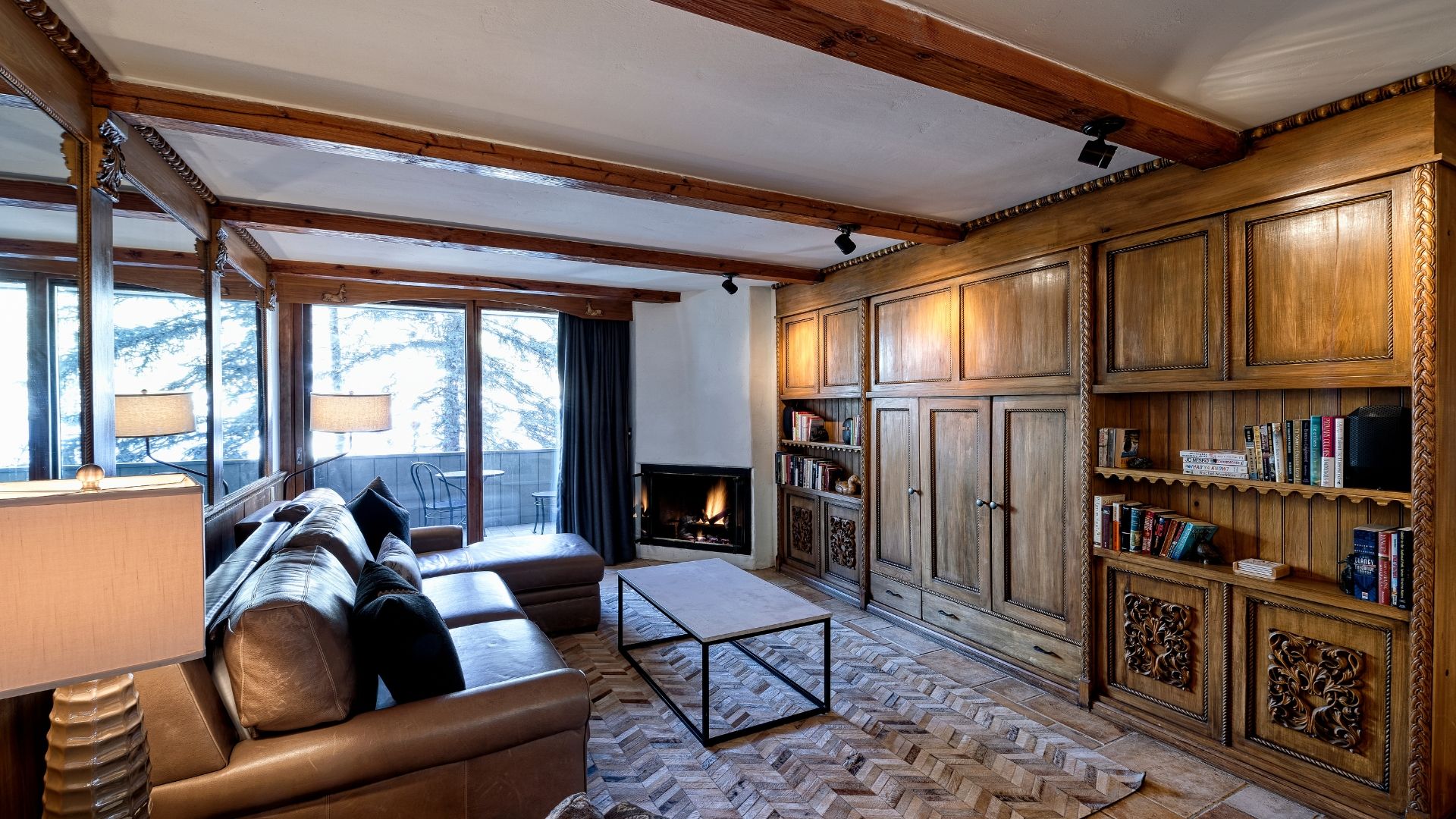 A cozy living room with a fireplace, leather sofa, wooden bookshelves, and a herringbone-patterned floor.