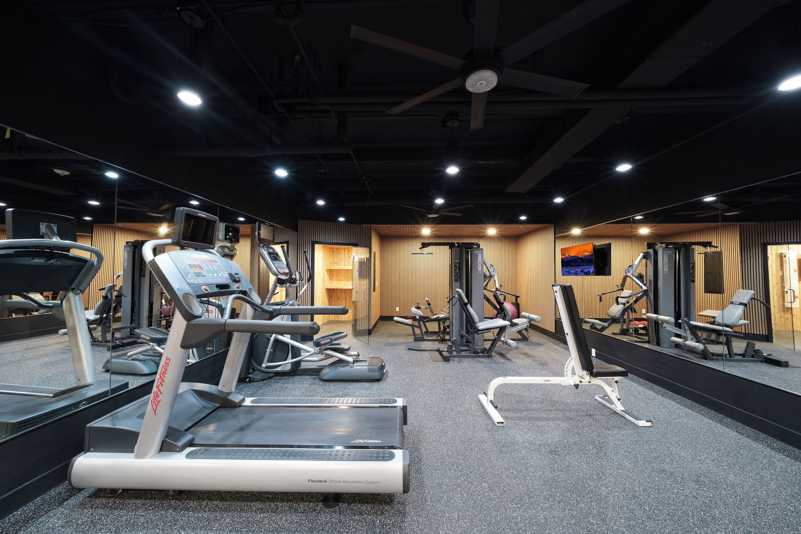 Un gimnasio con diversos equipos de ejercicio, incluyendo cintas de correr y máquinas de pesas, y un espejo grande que refleja el espacio.